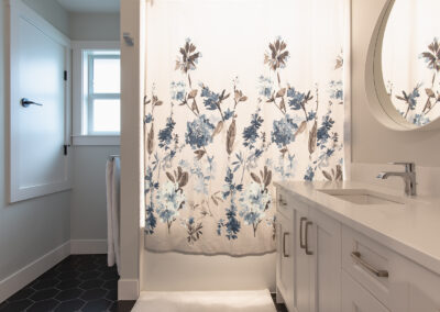 A bathroom with a floral shower curtain, white shaker cabinets, a round mirror, and a dark hexagon tile floor.