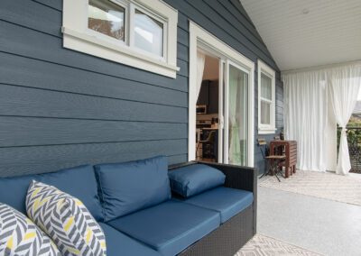 A dark wicker three-seat sofa with blue cushions and gray and yellow patterned pillows sits on a patio with dark blue siding, a sliding glass door, and sheer white curtains.