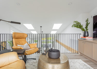 A modern loft space with a tan leather armchair featuring a pillow,a black metal railing, skylights, and a gray cabinet.