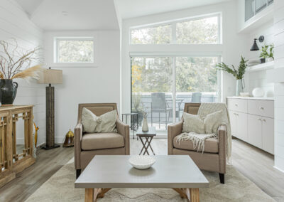 A modern living room with white shiplap walls, two beige armchairs, a gray coffee table, and a sliding glass door leading to a patio.