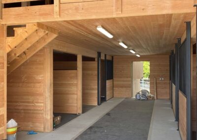 Interior of a wooden barn with stalls, a concrete aisle with a rubber mat, and a wooden loft area with railings.
