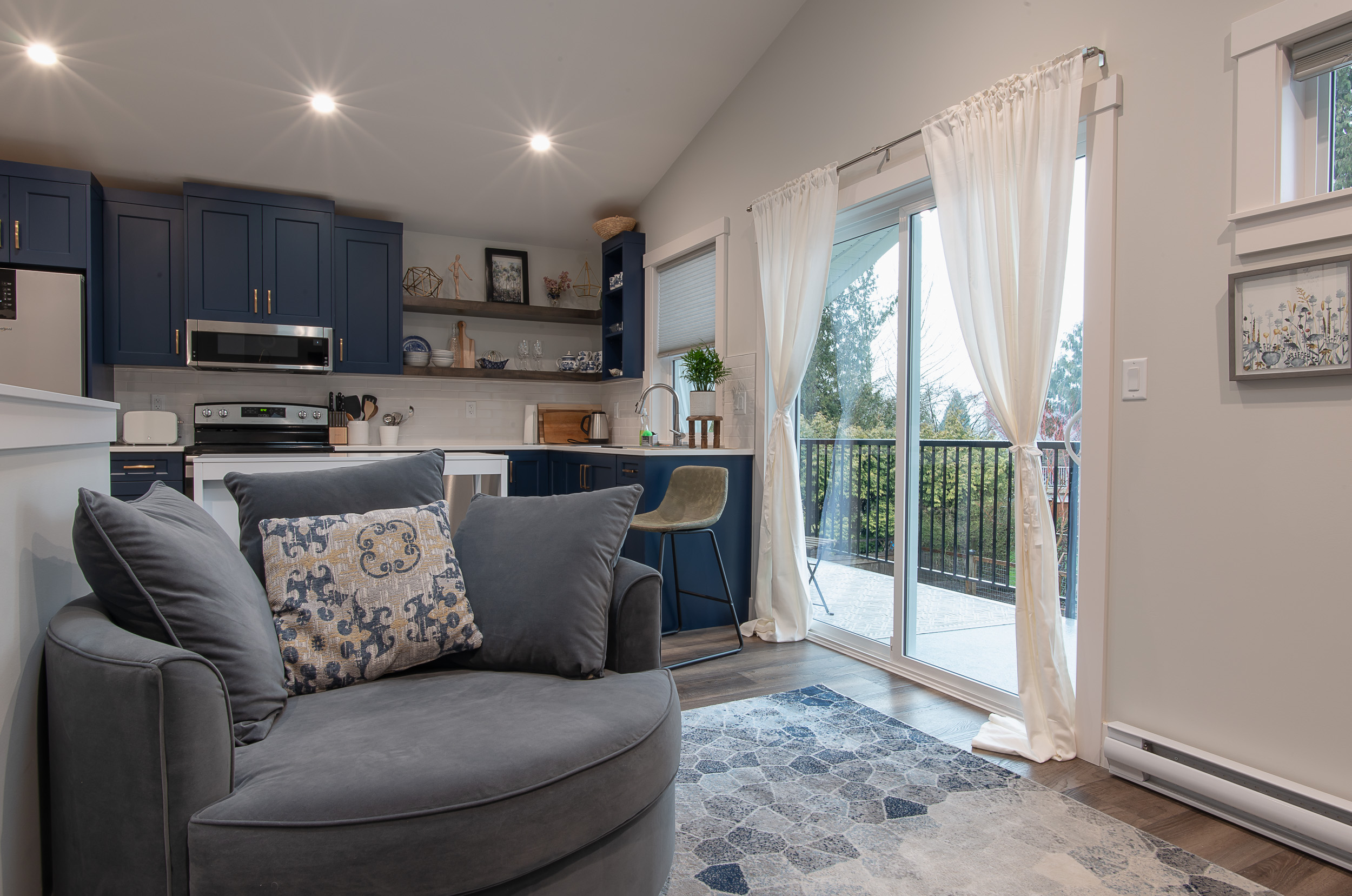 A large grey swivel armchair with patterned accent pillows sits on a blue geometric area rug in an open-concept living room and kitchen, highlighting the functional interior design of a multi-generational housing project in Langley by Bowline Construction.