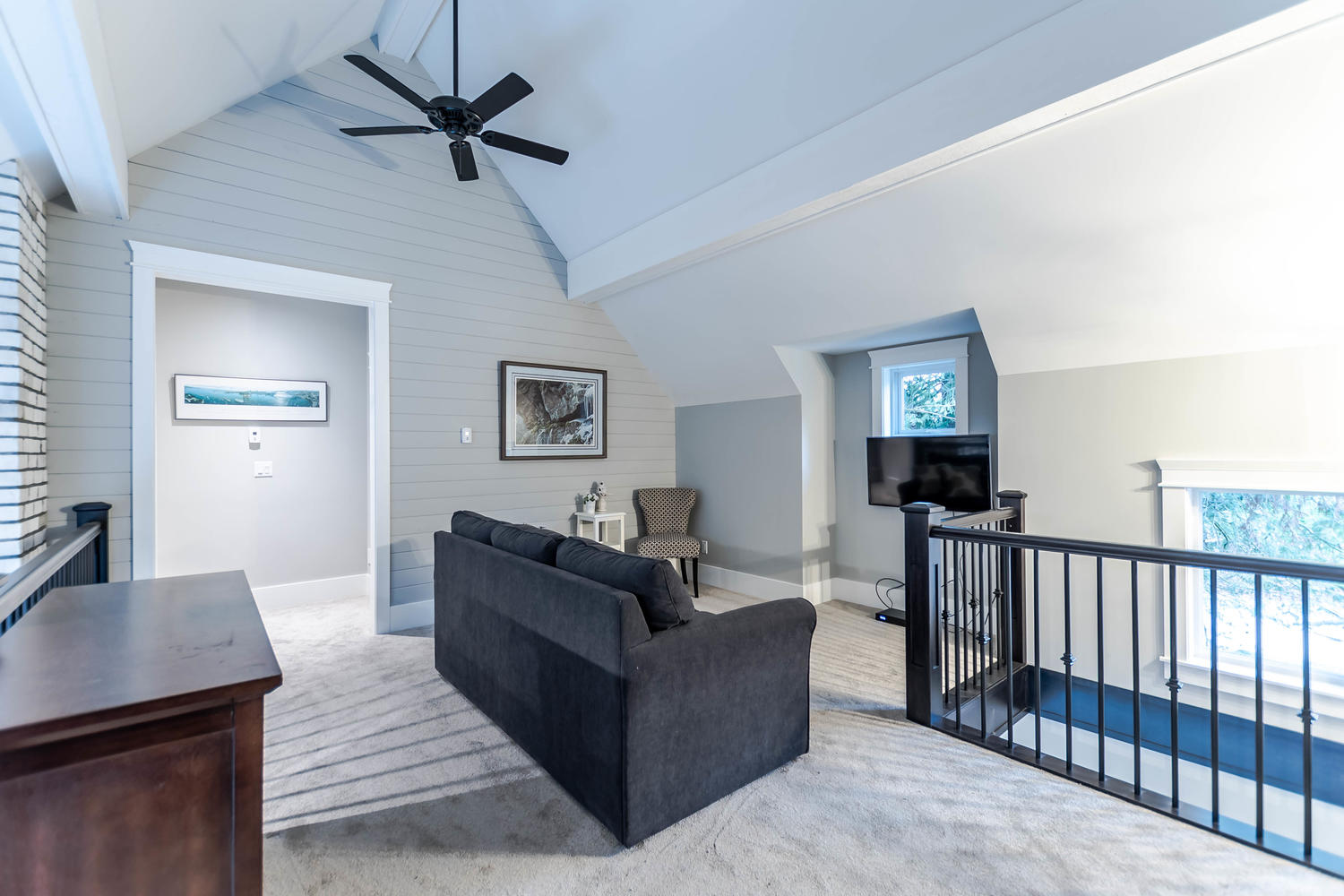 A bright second-story loft space features a dark grey sofa, light-colored carpeting, a vaulted ceiling with a black fan, and a protective black stair railing, demonstrating the versatile floor plans of a multi-generational housing project in Langley by Bowline Construction.