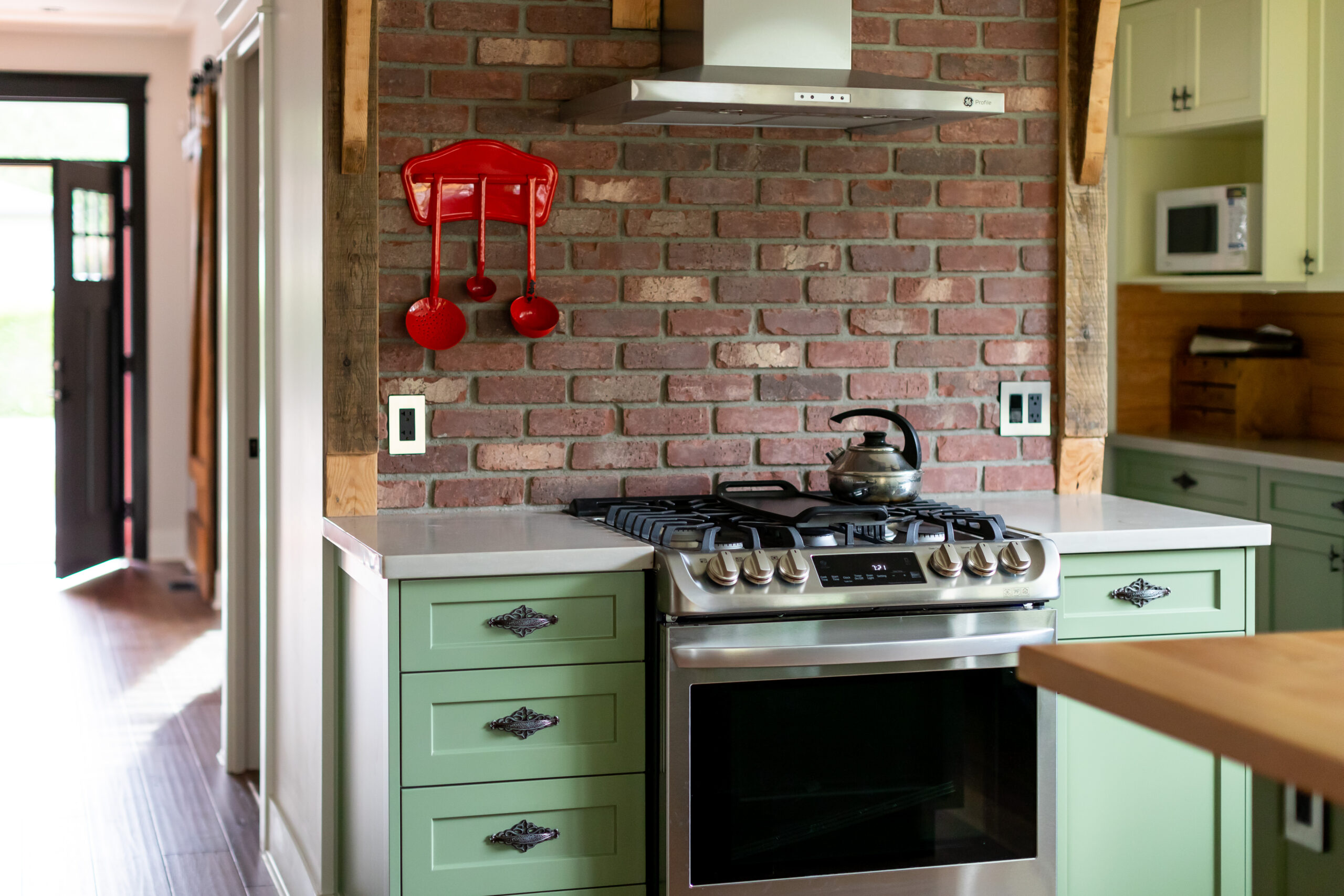 A stainless steel gas range and stainless range hood set against an exposed red brick accent wall with sage green cabinets, crafted by a kitchen contractor and kitchen renovation company in Langley City by Bowline Construction.